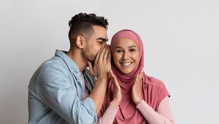 Pasangan suami istri Gossips Concept. Cheerful arab guy whispering something into his muslim girlfriends ear, millennial man sharing secret with islamic woman in hijab, having fun over grey studio background, free space