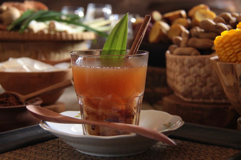 Bajigur, the traditional Sundanese hot herbal beverage of ginger tea, coffee, coconut milk and brown coconut sugar. In this variation, some palm fruit are added to the drink. The drink is garnished with pandanus leaf and cinnamon stick. It is arranged on a rustic wooden table together with typical steamed snacks that accompany this drink, such as steamed banana plantain, sweet potatoes, peanuts and corn on the cob.