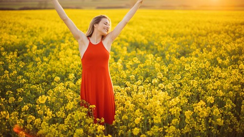 Beautiful young woman wearing bright red dress in yellow canola plant field. The field is spacious and the sun is showing in the background above the nearby hill. She is smiling and raising arms to the sky. Enjoying and celebrating freedom, happiness and positive thoughts and life.