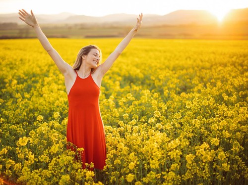 Beautiful young woman wearing bright red dress in yellow canola plant field. The field is spacious and the sun is showing in the background above the nearby hill. She is smiling and raising arms to the sky. Enjoying and celebrating freedom, happiness and positive thoughts and life.
