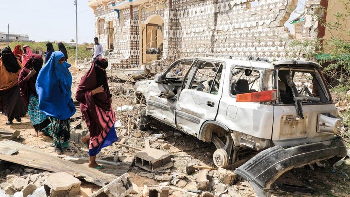 People walk next to a destroyed house and the wreckage of a car following an explosion provocked by Al-Shabaab militants during an attacke to a police station on the outskirts of Mogadishu, Somalia, on February 16, 2022. - Al-Shabaab militants launched simultaneous attacks on police stations around Somalias capital Mogadishu overnight, killing two girls and wounding over a dozen people, security officials said Wednesday. (Photo by Hassan Ali Elmi / AFP)