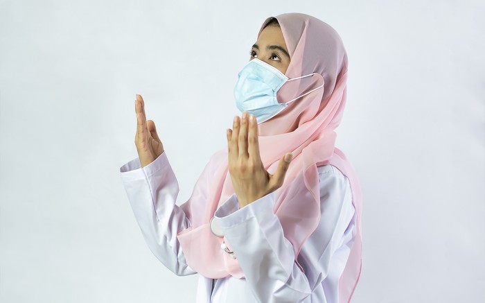 Ilustrasi wanita berhijab Muslim female doctor wearing headscarf, gown uniform and face mask to protect virus, standing and praying for hope on isolated white background. Medical, Science and Religion Concept
