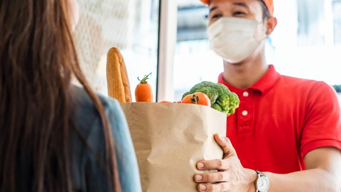 Asian deliver man wearing face mask in red uniform handling bag of food, fruit, vegetable give to female costumer in front of the house. Postman and express grocery delivery service during covid19.