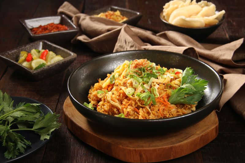 Nasi Goreng Jawa, the popular Indonesian dish of fried rice cooked Javanese style, using sweet soy sauce. The rice is mixed with shredded chicken and topped with omelet strips, scallion and fried shallot. Some accompanying condiments are arranged And a crumpled napkin are placed on the table.