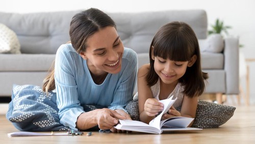 Caring smiling mother reading book with little kid girl lying on warm floor at home, mom or baby sitter playing having fun telling fairy tale to child daughter, underfloor heating, family activities