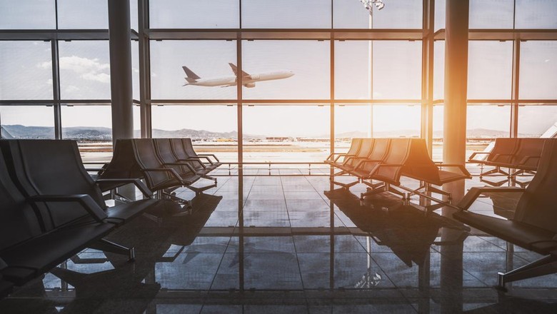 Wide-angle view of a modern aircraft gaining the altitude outside the glass window facade of a contemporary waiting hall with multiple rows of seats and reflections indoors of an airport terminal El Prat in Barcelona