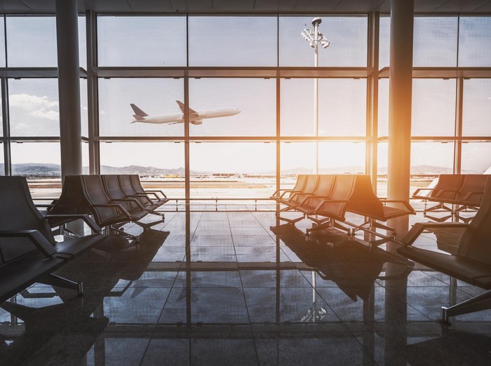 Wide-angle view of a modern aircraft gaining the altitude outside the glass window facade of a contemporary waiting hall with multiple rows of seats and reflections indoors of an airport terminal El Prat in Barcelona