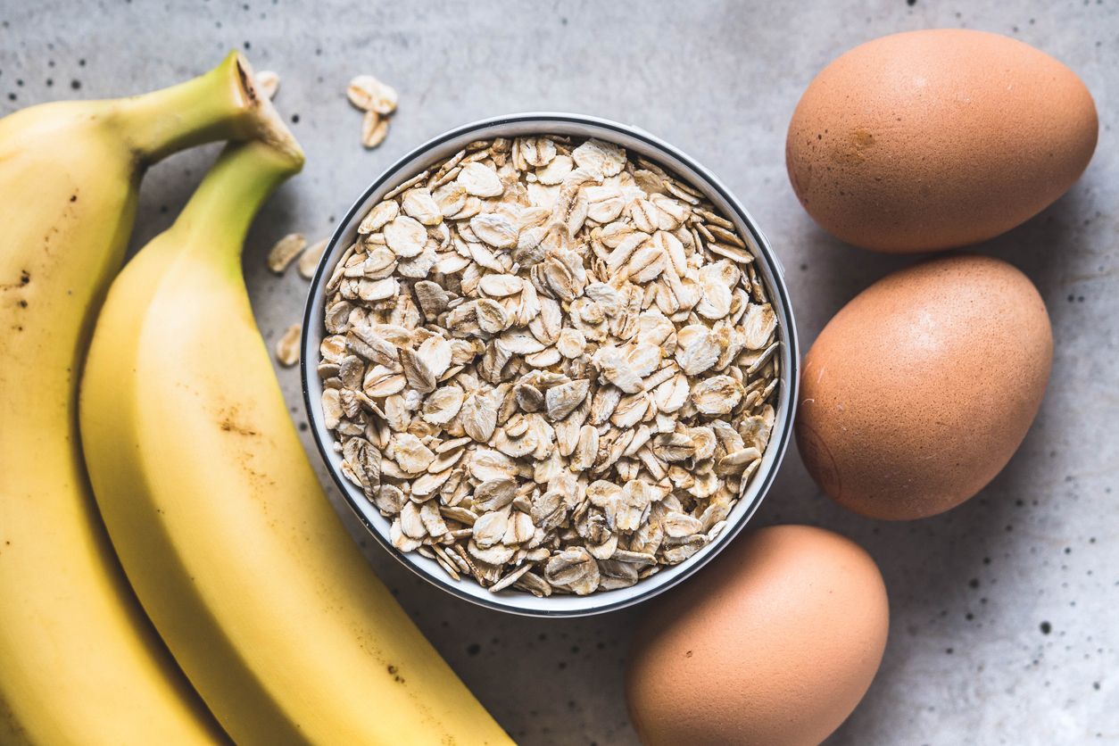 A bowl with oatmeal next to bananas and eggs on a concrete table. Healthy pancakes ingredients.