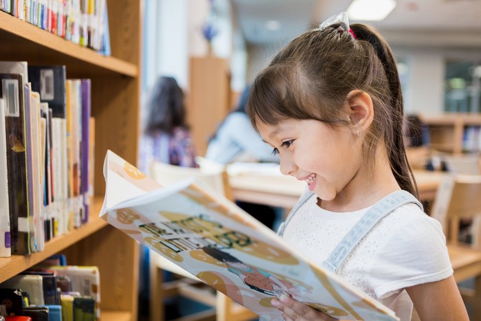 Adorable Hispanic schoolgirl smiles while delight as she reads a picture book while standing in the school library.
