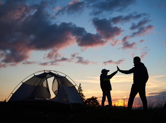Silhouette of man and child hikers give each other a high five near the tent at dawn against the backdrop of the mountains and the sun rising above them. Concept of healthy lifestyle.