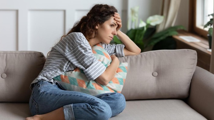Frustrated young lady sitting on sofa, cuddling pillow, looking away at window. Lost in thoughts unhappy stressed millennial woman regretting of wrong decision, spending time alone in living room.
