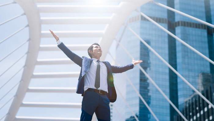 Celebrating success. Low angle view of excited young businessman keeping arms raised and expressing positive while standing outdoors with office building in the background