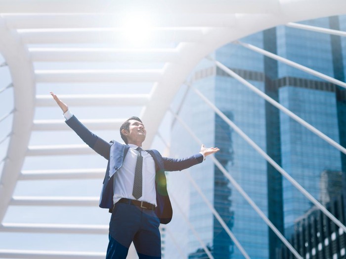 Celebrating success. Low angle view of excited young businessman keeping arms raised and expressing positive while standing outdoors with office building in the background