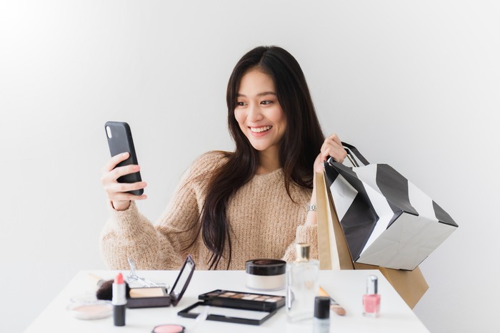 Ilustrasi wanita belanja kosmetik Asian beautiful women blogger are using the smartphone live streaming online with a shopping bag In the white room with cosmetics on table.Concept of online shopping business.