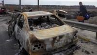 Seorang warga melewati mobil yang terbakar dalam tembakan Rusia saat ia melarikan diri dari kampung halamannya di jalan menuju Kyiv, di kota Irpin, barat laut Kyiv, Sabtu, (12/3/2022). (AP Photo/Efrem Lukatsky