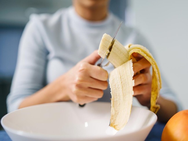 Caucasian woman sitting by the table at home cutting banana - Adult girl female preparing fruit salad at home - healthy eating concept copy space close up