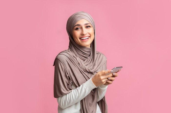 Cool application. Happy muslim girl in hijab using smartphone, texting or browsing internet and looking at camera, pink background with free space