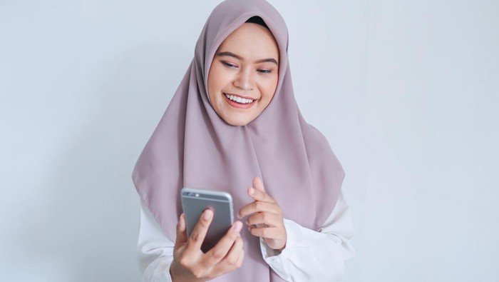 Young Asian Islam woman wearing headscarf is smile and happy in what she see on the smartphone. Indonesian woman on gray background.