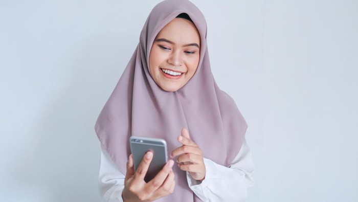 Young Asian Islam woman wearing headscarf is smile and happy in what she see on the smartphone. Indonesian woman on gray background.