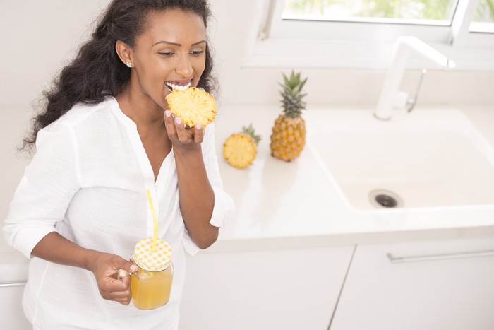 Happy woman eating pineapple and drinking freshly squeezed pineapple juice. Attractive Ethiopian woman wearing a white shirt, standing, leaning on the kitchen countertop.