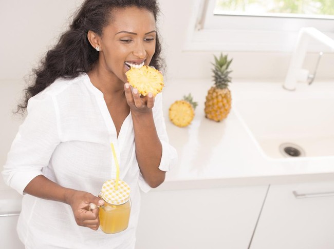 Happy woman eating pineapple and drinking freshly squeezed pineapple juice. Attractive Ethiopian woman wearing a white shirt, standing, leaning on the kitchen countertop.