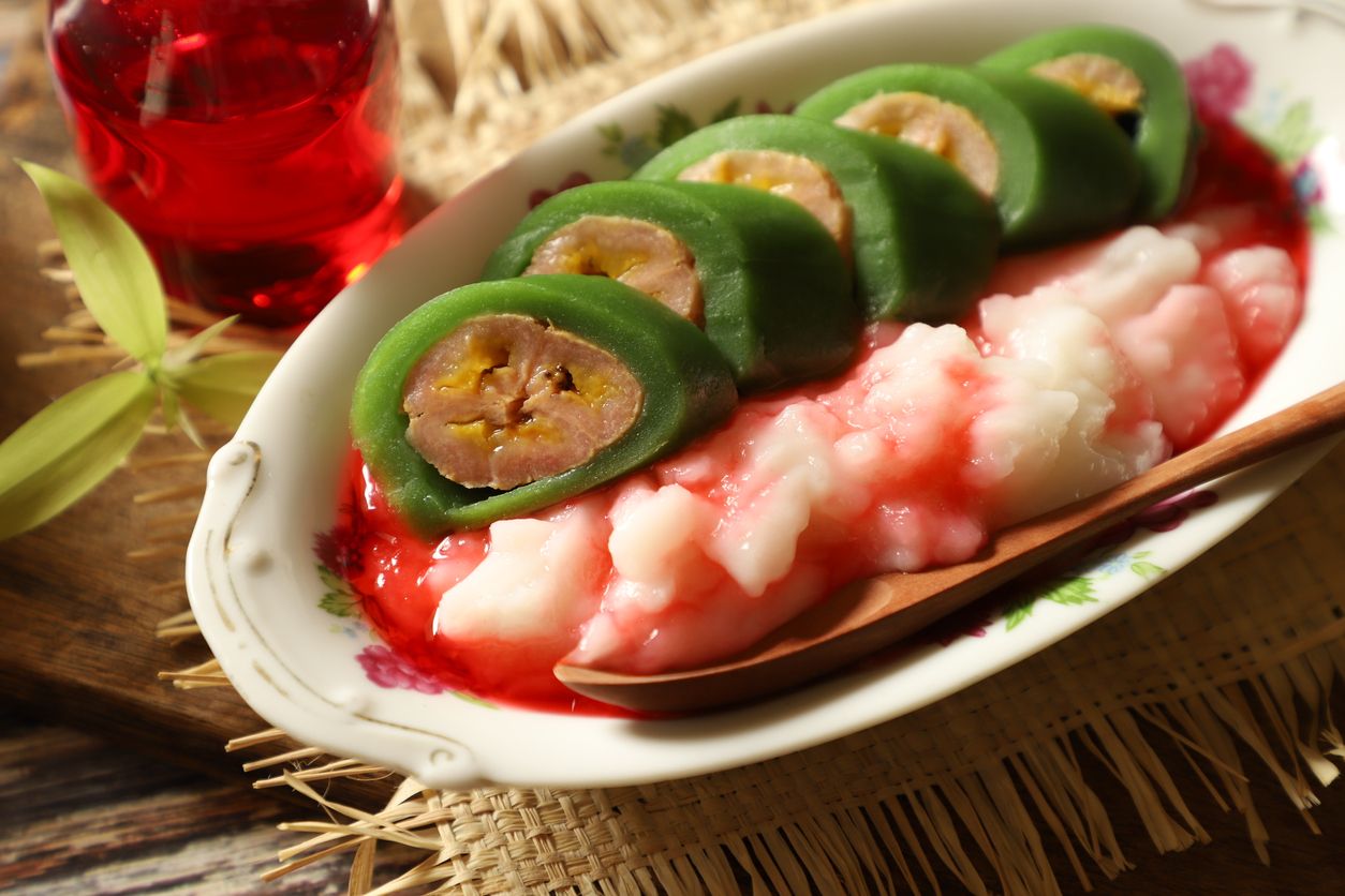 Es Pisang Ijo, the popular dessert of plantain banana roll with coconut milk custard and red sugar syrup from Makassar (Ujung Pandang), South Sulawesi. The banana is wrapped with pandan crêpe; then served in slices on the custard. The dessert is plated on an vintage oval plate and placed on straw mat. An extra syrup in a small jug is placed next to the dessert.