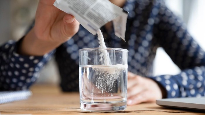 1257836796 Close up young indian woman pouring soluble anti-influenza powder in glass of water. Sick millennial girl student feeling unwell, relieving grippe fever flu symptoms, high temperature, headache.