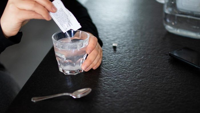 1257836796 Close up young indian woman pouring soluble anti-influenza powder in glass of water. Sick millennial girl student feeling unwell, relieving grippe fever flu symptoms, high temperature, headache.