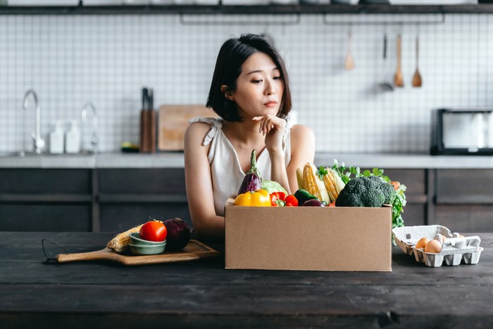 Young Asian woman received a box full of colourful and fresh organic groceries ordered online by home doorstep delivery. She is planning to cook a healthy meal in the kitchen at home. Healthy eating lifestyle