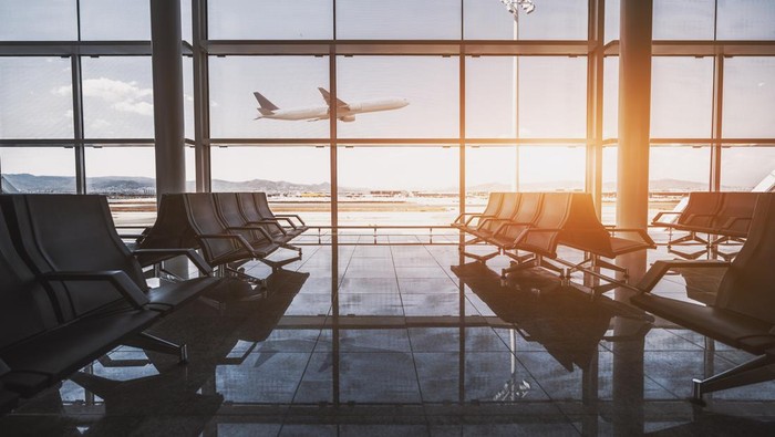 Wide-angle view of a modern aircraft gaining the altitude outside the glass window facade of a contemporary waiting hall with multiple rows of seats and reflections indoors of an airport terminal El Prat in Barcelona