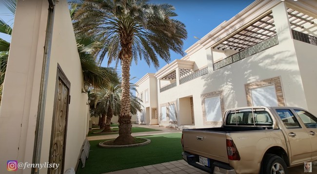 Halaman rumah terlihat luas dan juga asri. Ditumbuhi dengan pohon-pohon kurma yang cukup besar, rumah tersebut jadi tampak rindang dan sejuk. Dominasi warna coklat pada desain bangunan rumah juga membuatnya semakin sedap dipandang. Foto: YouTube/Fenny Listiana