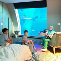 Pemandangan bawah laut itu langsung menghadap ke tempat tidur di dalam kamar hotel. Di sampingnya juga tampak disediakan meja dan kursi untuk duduk dan menikmati pemandangan akuarium bawah laut. Terlihat ibu Titi Kamal dan anak-anaknya begitu senang menghabiskan waktu di dalam kamar hotel tersebut. Foto: Instagram/@titi_kamall