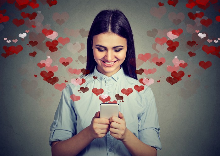 661806170 Beautiful happy woman sending love text message on mobile phone with red hearts flying away from screen isolated on gray background. Positive human emotions face expressions