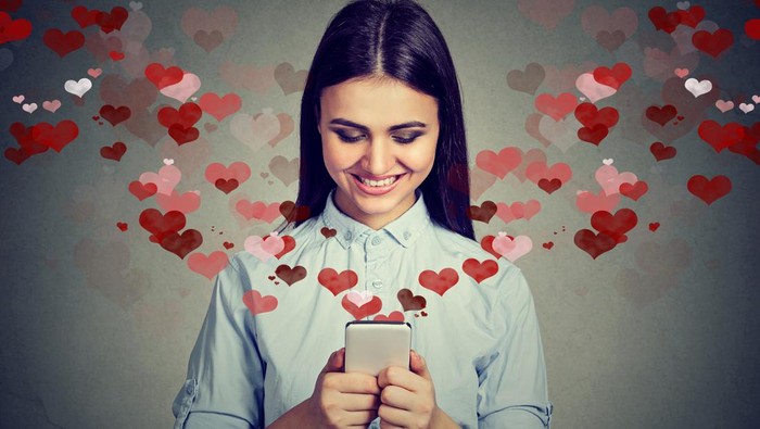 Beautiful happy woman sending love text message on mobile phone with red hearts flying away from screen isolated on gray background. Positive human emotions face expressions