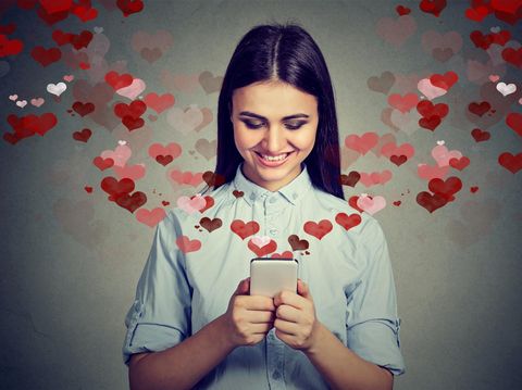 Beautiful happy woman sending love text message on mobile phone with red hearts flying away from screen isolated on gray background. Positive human emotions face expressions