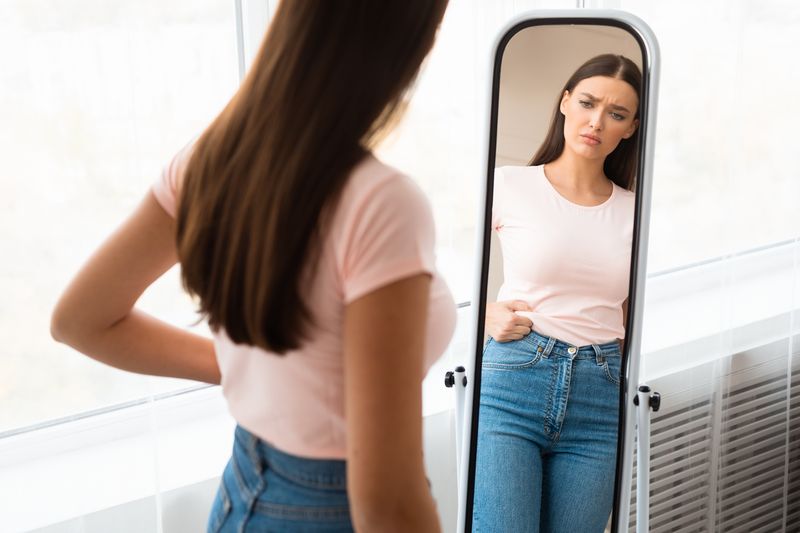 Weight Gain. Unhappy Girl Touching Drooping Belly Standing In Front Of Mirror At Home. Empty Space, Selective Focus