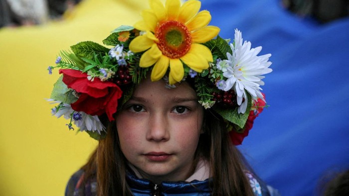 A young girl looks at a camera as people carry a huge Ukrainian flag as they participate in a peaceful demonstration 