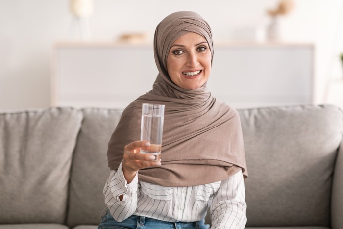 Happy Middle-Aged Muslim Woman In Hijab Posing Holding Glass Of Water Drinking Healthy Liquid For Hydration Sitting On Couch At Home, Smiling To Camera. Stay Hydrated Concept