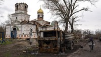Seorang wanita dengan sepeda mengendarai kendaraan yang terbakar, di depan Gereja yang rusak di Lukashivka, dekat kota Chernihiv di Ukraina utara, pada hari Jumat, (22/4/2022). Sebuah salib logam tetap berada di dalam gereja Ortodoks dari batu bata yang hancur dan batu menghitam. Penduduk mengatakan tentara Rusia menggunakan rumah ibadah untuk menyimpan amunisi, dan pasukan Ukraina menembaki gedung itu untuk membuat Rusia pergi. (AP Photo/Petros Giannakouris)