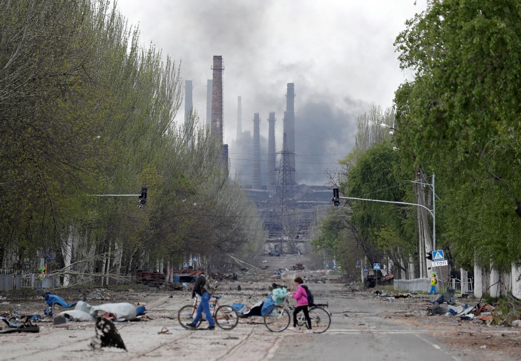 Orang-orang berjalan dengan sepeda mereka di seberang jalan saat asap membubung di atas pabrik Azovstal Iron and Steel Works selama konflik Ukraina-Rusia di kota pelabuhan selatan Mariupol, Ukraina, Senin (2/5/2022). (REUTERS/Alexander Ermochenko)