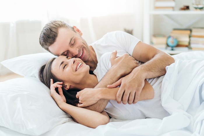 Close up of young couple lying in bed together at the sunny morning. Man in woman in love smiling to each other and giving hugs.