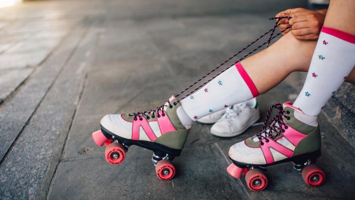 sepatu roda Girl is sitting on the floor and tying laces on her right roller. She is pulling them back. Also she put her right feet on the back rollers wheels