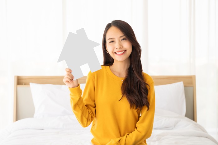 Ilustrasi Wanita Jual Rumah Beautiful asian woman happy and smile sitting on bed in bedroom and holding paper house symbol
