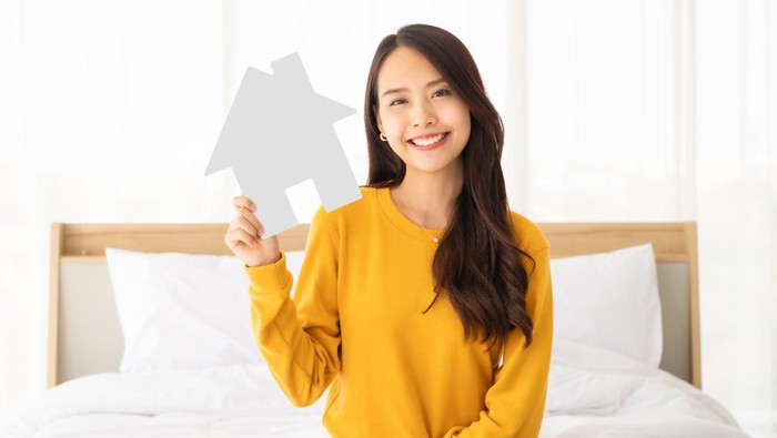 Beautiful asian woman happy and smile sitting on bed in bedroom and holding paper house symbol