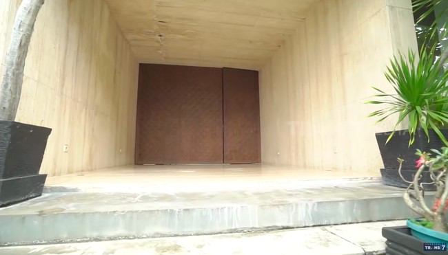 Anti-mainstrem, pintu rumah Devi selain besar, tak ada gagang yang menempel. Satu-satunya cara untuk membuka pintu kayu yang berat ini adalah dengan mendorong. Sebagai informasi, harga pintunya mencapai Rp 25 juta.   (Foto: YouTube/Trans7 Official)