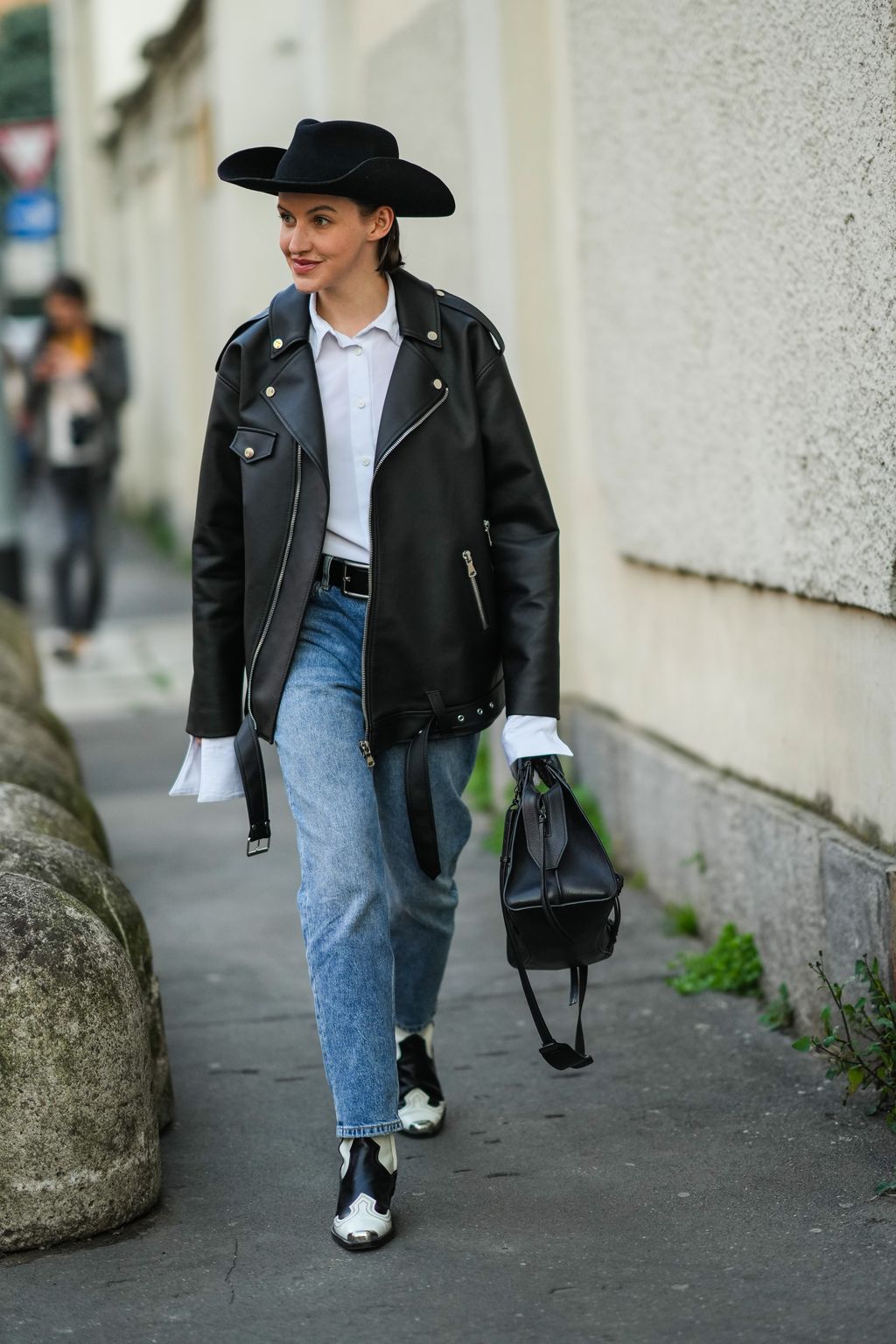 MILAN, ITALY - FEBRUARY 23: A guest wears a black felt hat, a white shirt, a black shiny leather oversized zipper jacket, blue denim cigarette jeans pants, a black shiny leather handbag from Balenciaga, black and white leather yoke and metallic pointed block heels ankle boot, outside the Alberta Ferretti fashion show, during the Milan Fashion Week Fall/Winter 2022/2023 on February 23, 2022 in Milan, Italy. (Photo by Edward Berthelot/Getty Images)