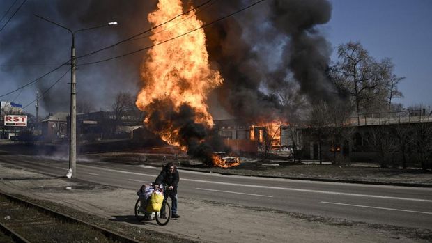 Seorang pria melarikan diri dengan barang-barangnya saat api melalap kendaraan dan bangunan setelah tembakan artileri Rusia pada hari ke-30 serangannya ke Ukraina di kota timur laut Kharkiv, Ukraina (25/3/2022). Serangan Rusia menargetkan fasilitas medis di Ukraina kota kedua Kharkiv pada 25 Maret 2022, menewaskan sedikitnya empat warga sipil dan melukai beberapa lainnya, kata pejabat Ukraina. (Photo by Aris Messinis / AFP)