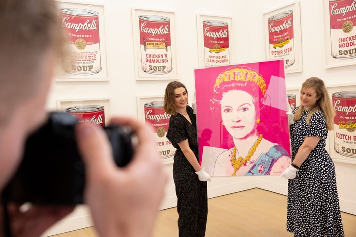 LONDON, ENGLAND - JUNE 01: Gallery staff pose with a portrait of Queen Elizabeth II by artist Andy Warhol at Phillips auction house on June 01, 2022 in London, England. Phillips estimates the print to sell for £100,000 – 150,000 at auction, timed to coincide with the Queen's platinum jubilee. The portrait, part of Warhol's 1985 