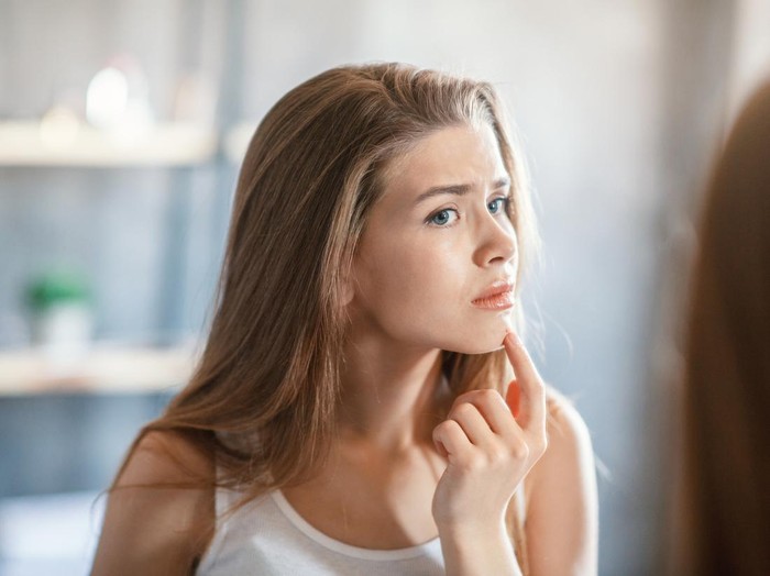 Upset millennial girl touching pimple on her chin in front of mirror at home