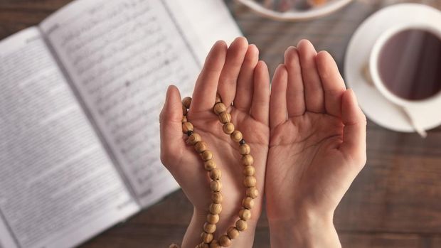 female hand of prayer with wooden beads in sunlight, iftar concept, Ramadan month, Koran, plate of dried fruit, Cup of tea on wooden table
